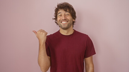 A cheerful young hispanic man with a beard pointing at something over an isolated pink background,...