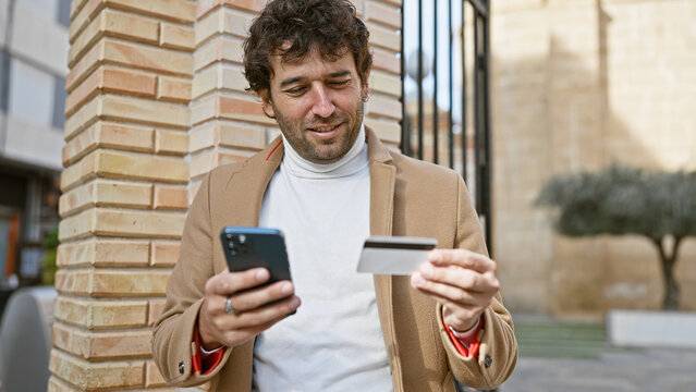 Hispanic man smiling at phone holding credit card, urban brick building background.