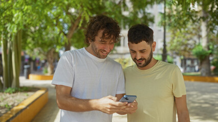 Men smiling while looking at a smartphone in an outdoor urban park, surrounded by greenery and...