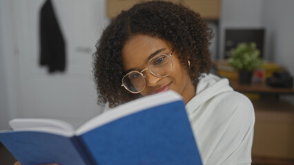 A beautiful young african american woman with curly hair reading a blue book in an indoor office...