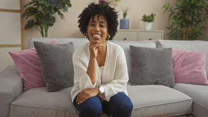 Smiling african american woman with curly hair sitting on a cozy sofa in an elegantly decorated living room at home.