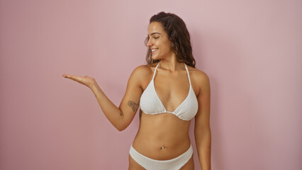Fototapeta premium Young woman in white bikini presenting something with extended arm against pink background