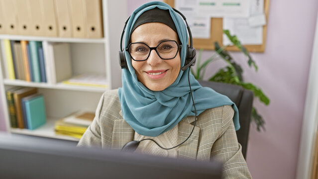 Smiling mature woman in hijab and headset at workplace with bookshelf and plant background.