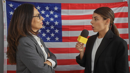 Two professional women, reporter with microphone interviewing a leader in front of an american flag indoors.