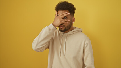 African american man in hoodie standing against yellow background covering eyes with hand