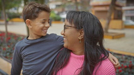 A smiling woman hugs a joyful boy in a sunny park, showcasing a warm family moment between mother and son outdoors.