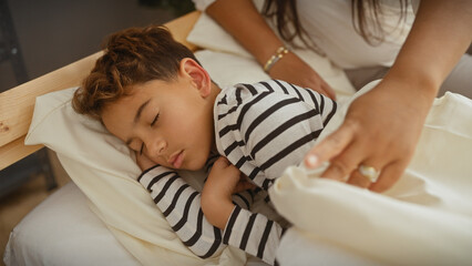 A young boy sleeps peacefully on a bed while a woman gently tucks him in, depicting a tender family moment indoors.