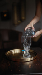 A person pouring water into a glass on a gold tray set on a wooden table, evoking a sense of hospitality or service.