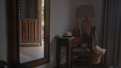 A serene interior scene featuring a panama hat, wooden lamp, console table, cushioned chair, glass bottle, rustic door, and a hanging straw bag by the curtains.