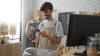 Young hispanic man with a beard smiling while using a smartphone in a bakery shop interior with...