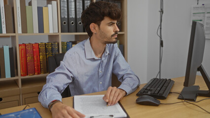 Handsome young man working indoors in an office setting with shelves of books and folders, wearing a striped shirt, focusing on documents at a desk with a computer