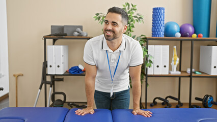 A smiling hispanic man with a beard wearing a lanyard in a physical therapy clinic