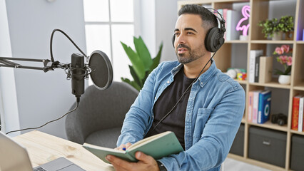 Fototapeta premium Hispanic man with beard sitting in radio studio wearing headphones holding a book.