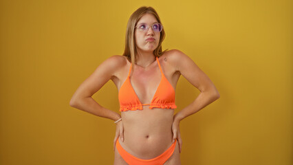 Obraz premium Young blonde woman posing against a yellow background in an orange bikini looking thoughtful with hands on hips