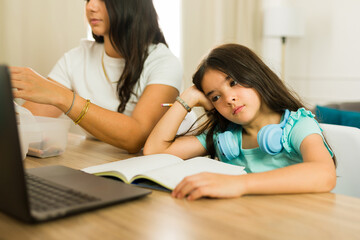 Unmotivated elementary school girl feeling bored while studying with her mother and looking sad at the kitchen table