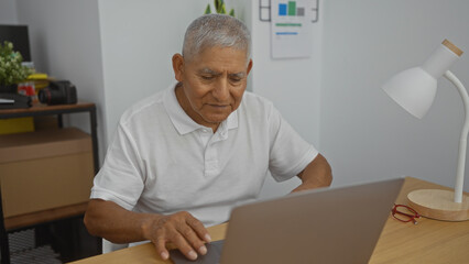 Mature, hispanic, grey-haired man working on a laptop in an office with a focused expression, desk lamp, and office supplies.