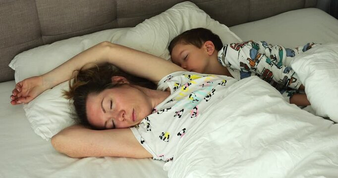 Tired mother and little son sleep together on soft double bed in early morning. Young female with young boy enjoy calm rest in bedroom. Family bonding