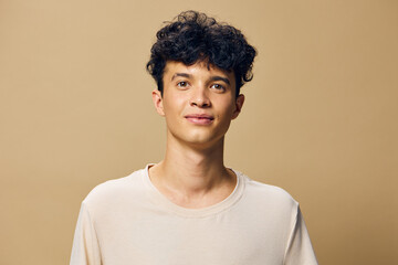 Young man with curly hair smiling against a warm beige background, wearing a light t shirt The simple and neutral setting highlights his cheerful expression and youthful energy