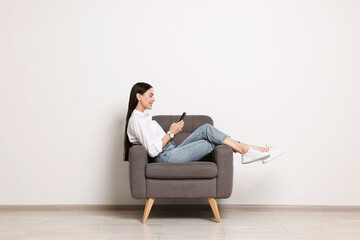 Smiling woman with smartphone sitting on armchair against white wall