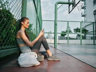 Young woman sitting by a basketball court checking her phone, wearing activewear and earbuds, with green fencing and urban background