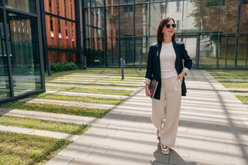 A stylish businesswoman confidently walks outdoors in a modern office complex during the day