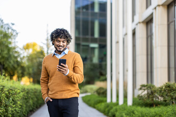 Young man in orange sweater uses phone while walking in urban area. Happy with technology interaction. Sunny day highlights blend of nature and architecture in modern city setting.