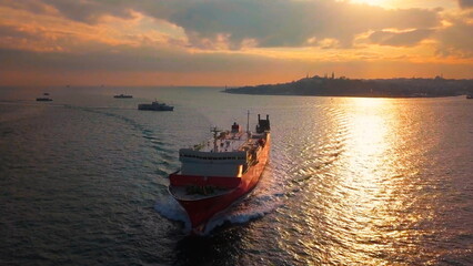 Naklejka premium A car carrier navigate Sarayburnu offshore at sunset. Iconic mosques silhouetted against dramatic sky. Bosphorus, Turkey