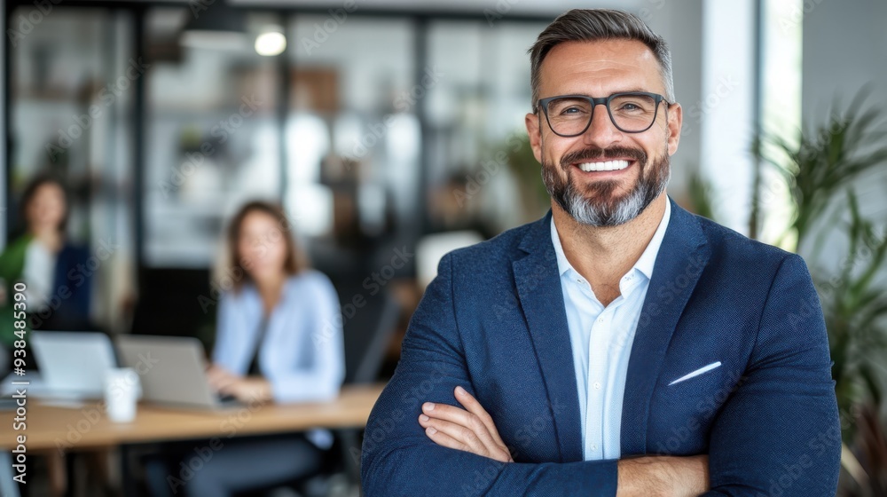 Wall mural A smiling man with a beard, styled hair, and glasses, wearing a blue blazer, stands confidently in a modern office environment, exuding professionalism and poise.