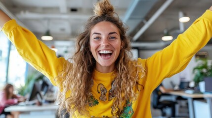 Fototapeta premium A joyful woman in a vibrant yellow sweater celebrates success with raised arms in a bright and modern office space, showcasing enthusiasm and a sense of achievement.