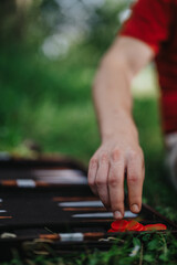 A person in a red shirt is playing backgammon on the grass. The image highlights the hand and red game pieces, evoking a sense of leisure and strategy.