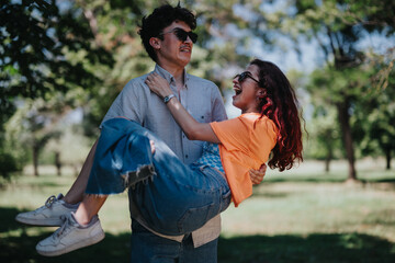 A joyful couple shares a playful moment in a park. The man lifts the woman, both smiling and wearing sunglasses, enjoying a sunny day together.