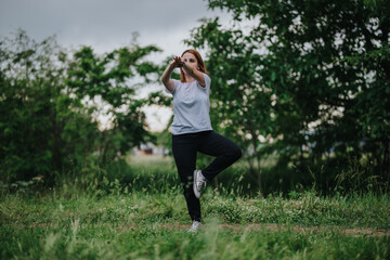 A woman performs a yoga pose in a tranquil park surrounded by lush greenery, embodying peace,...