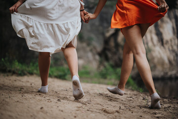 Two women holding hands and walking barefoot on a dirt path surrounded by nature, showcasing freedom and friendship.