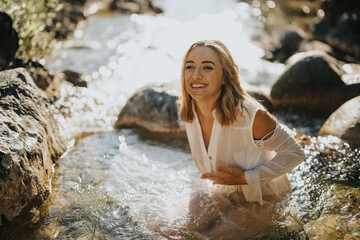 Happy young woman in a white dress smiling while sitting in a sunlit stream. Natural beauty enjoying outdoors in nature.