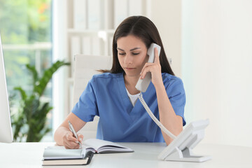 Beautiful nurse working with clients by telephone at table in hospital office