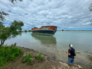 A fisherman is fishing on the shore of Lake Ontario against the background of an old abandoned ship