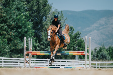 Equestrian rider and horse jumping over a colorful hurdle outdoors with scenic mountains and trees in the background on a sunny day.