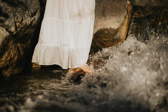 Close-up of a woman in a white dress standing with bare feet in a flowing stream near rocks, evoking calm and tranquility.
