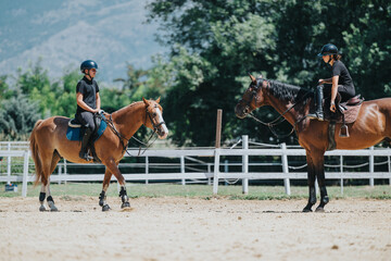 Two individuals riding horses in a sunny outdoor arena, wearing helmets and equestrian gear,...