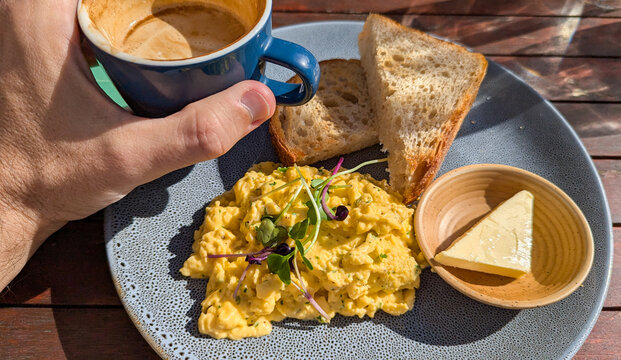 Eating a delicious breakfast of scrambled eggs with micro greens, toast and butter with a golden flat white coffee on wooden table