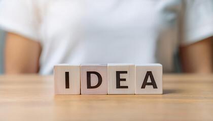 Naklejka premium 木製のブロックに「IDEA」という文字が書かれており、それがテーブルの上に並べられています。背景には白いシャツを着た人物がぼんやりと写っており、アイデアの概念や創造性を表現しています。全体的にシンプルでフォーカスが「IDEA」に当てられています。generative AI