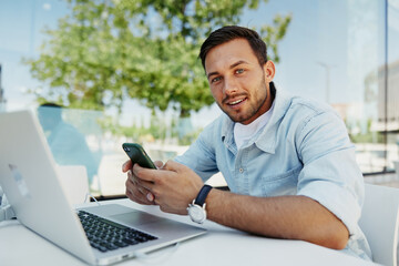 Young man working remotely outdoors, smiling while holding a smartphone and using a laptop