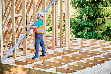Fototapeta premium Carpenter constructing two-storey wooden frame house. Man inspects walls for levelness using spirit level, wearing protective overalls, helmet and orange vest. Concept of eco-friendly construction.