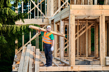 Carpenter constructing wooden-framed house near the woods. Bearded man with spectacles holding large joist, wearing work clothes and helmet. Concept of modern and eco-friendly building.