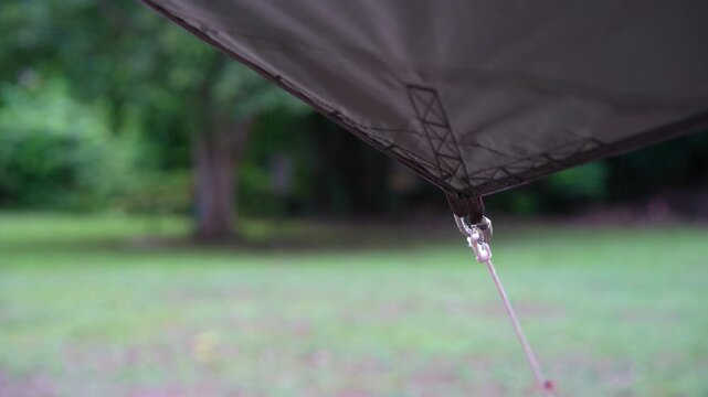 camping on rainy season with closeup rain water drips droplets to tip tarp and flow motion on fly sheet tent with carabiner paracord rope for holiday vacation on nature forest garden with rain sound