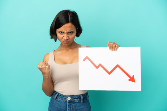 Young mixed race woman isolated on blue background holding a sign with a decreasing statistics arrow symbol and angry