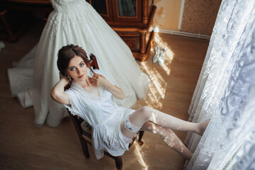 A woman in a white dress is sitting on a chair in front of a window. She is wearing white stockings and a white gown