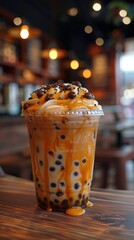 Caramel bubble tea topped with whipped cream, chocolate pearls, and caramel drizzle in a plastic cup on a wooden table