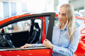 Smiling caucasian woman hold door of new red car on background of showroom