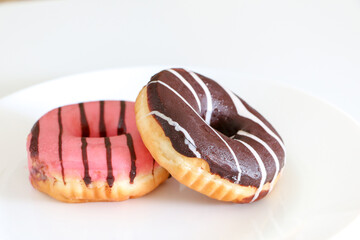 Two doughnut (donut) rings glazed and striped with strawberry pink, chocolate brown and white sugar icing, on white dish. Moist just out of refrigerator. Close up of sweet sugary but unhealthy food